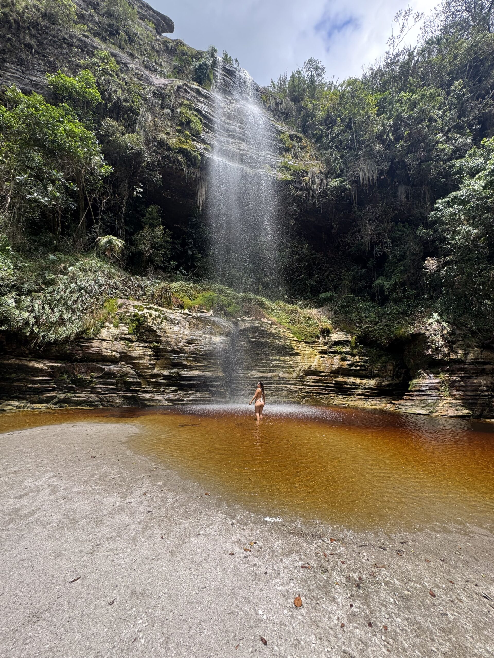 Cachoeira em Ibitipoca, Minas Gerais, com água caindo suavemente sobre paredões de pedra e formando uma piscina natural de tons dourados.