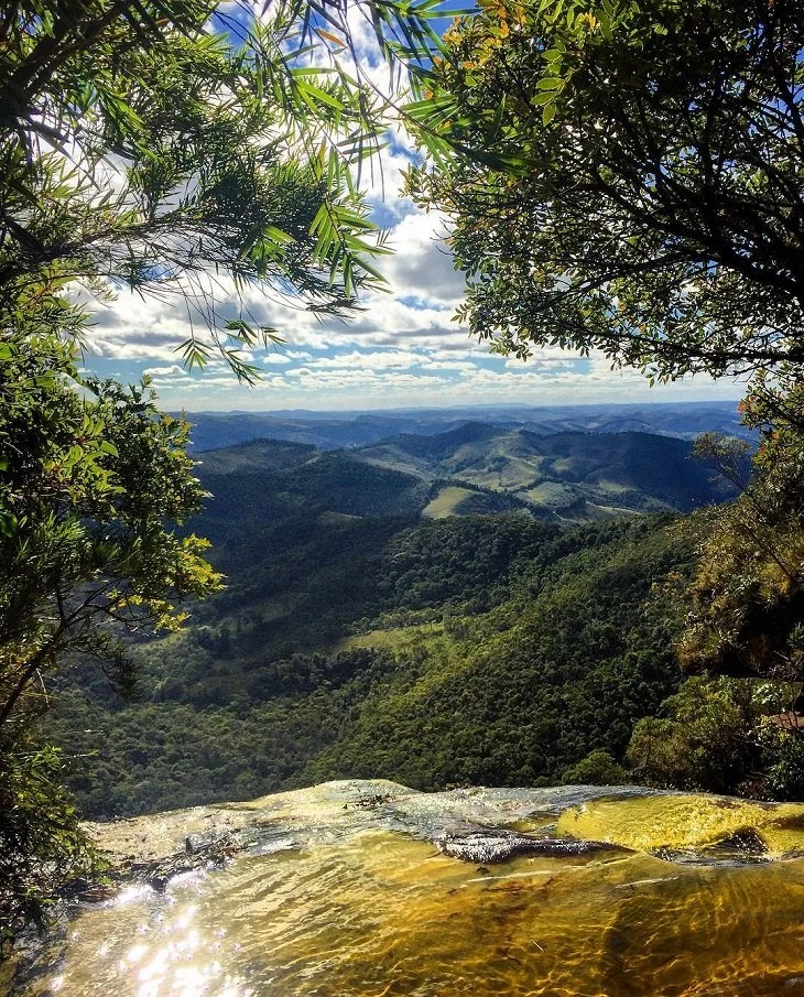 Vista panorâmica das montanhas de Ibitipoca a partir da Janela do Céu, com vegetação verde, céu azul e a água dourada da cachoeira refletindo a luz do sol.